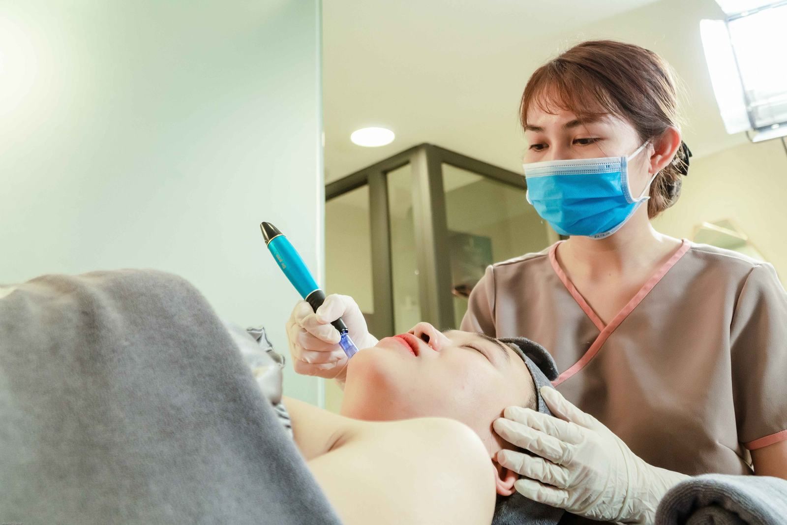 A masked provider holding a pen-style skin treatment device near a client's chin during a facial skin treatment