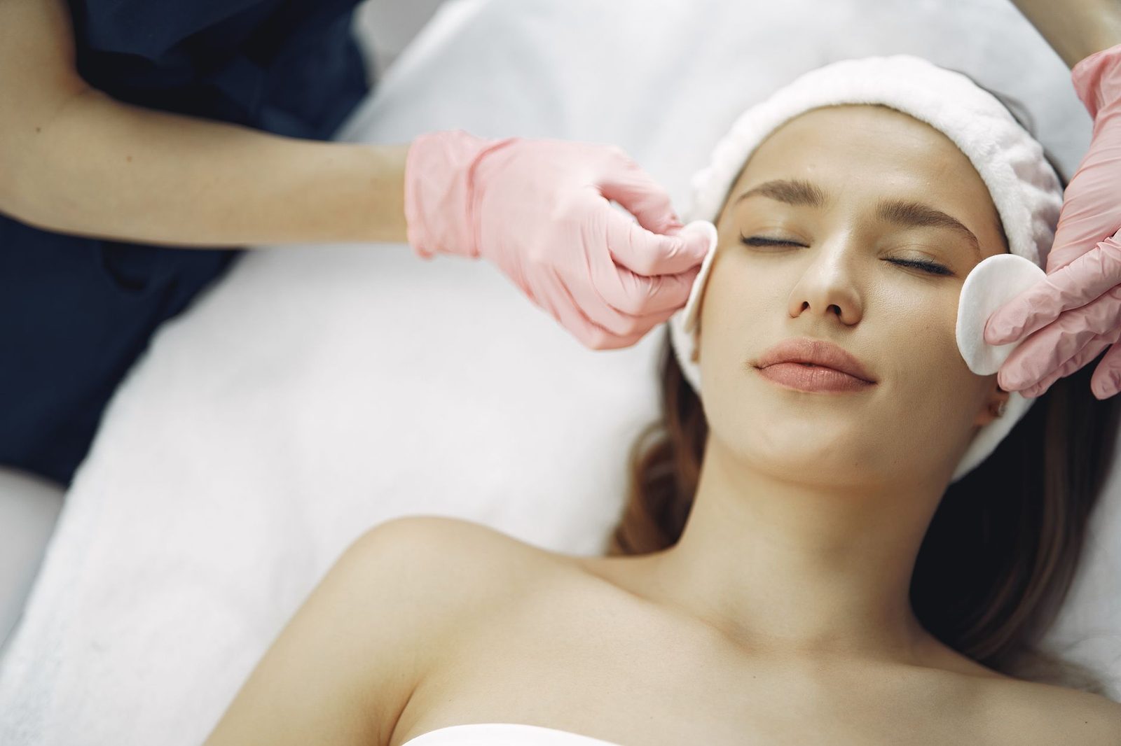 A woman lying back in a headband while pink-gloved hands wipe her face with cotton pads during a facial